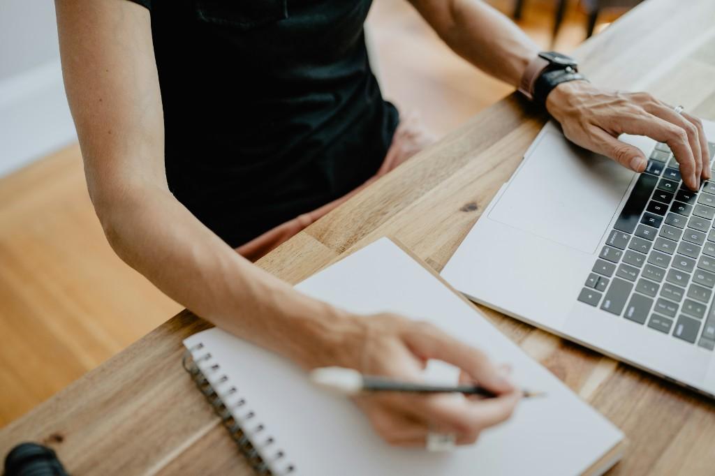 Person taking notes in a notebook beside a laptop while preparing research